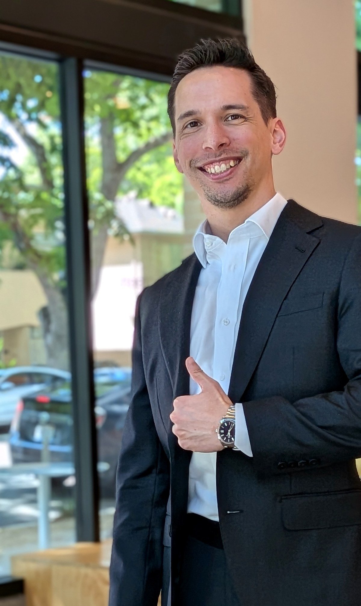 Josh Croft in a charcoal suit with open collar, giving a thumbs-up